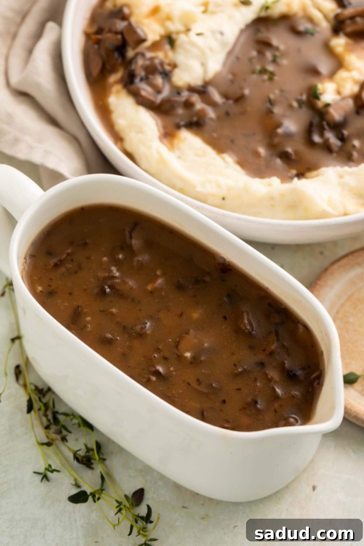 Irresistible Vegan Mushroom Gravy 3 A large ceramic white gravy boat holding rich brown vegan mushroom gravy, next to a bowl of mashed potatoes generously topped with the gravy.