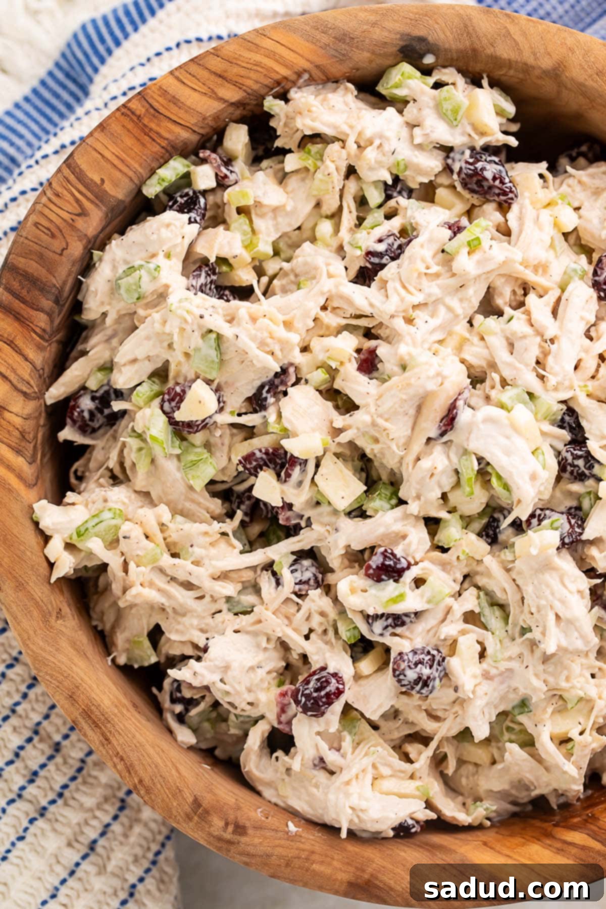A large wooden bowl of creamy cranberry chicken salad, adorned with fresh parsley, sits invitingly next to a blue and white kitchen towel, ready to be served.