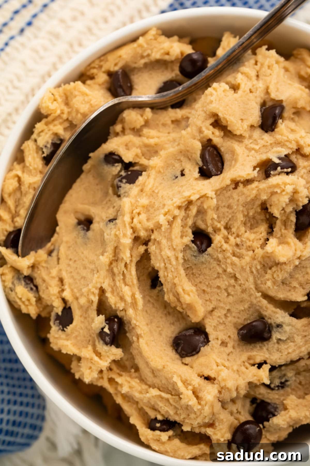 Close-up of a bowl of edible vegan cookie dough with a spoon resting in the dough, showcasing its creamy texture and chocolate chips.