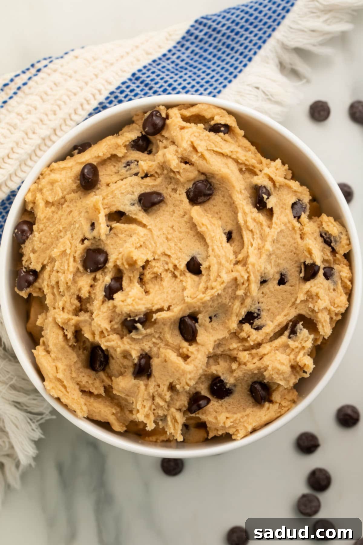 A bowl of vegan cookie dough rests on a kitchen counter, surrounded by loose chocolate chips and a blue and white striped kitchen towel, creating an inviting scene.