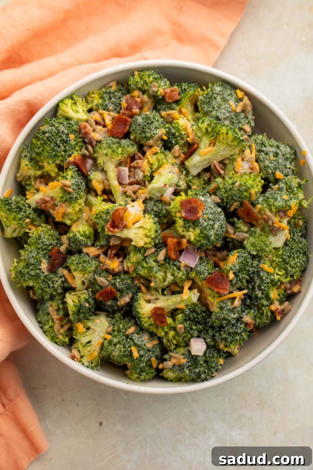 On a neutral countertop, next to a light peace cloth napkin, sits a large white bowl filled with a keto salad made with chopped broccoli florets, bacon, cheddar cheese, red onion, and a light mayo dressing.