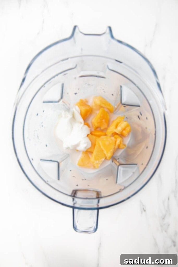 Cubed mango, pineapple, almond milk, and Greek yogurt inside a blender, ready to be mixed.