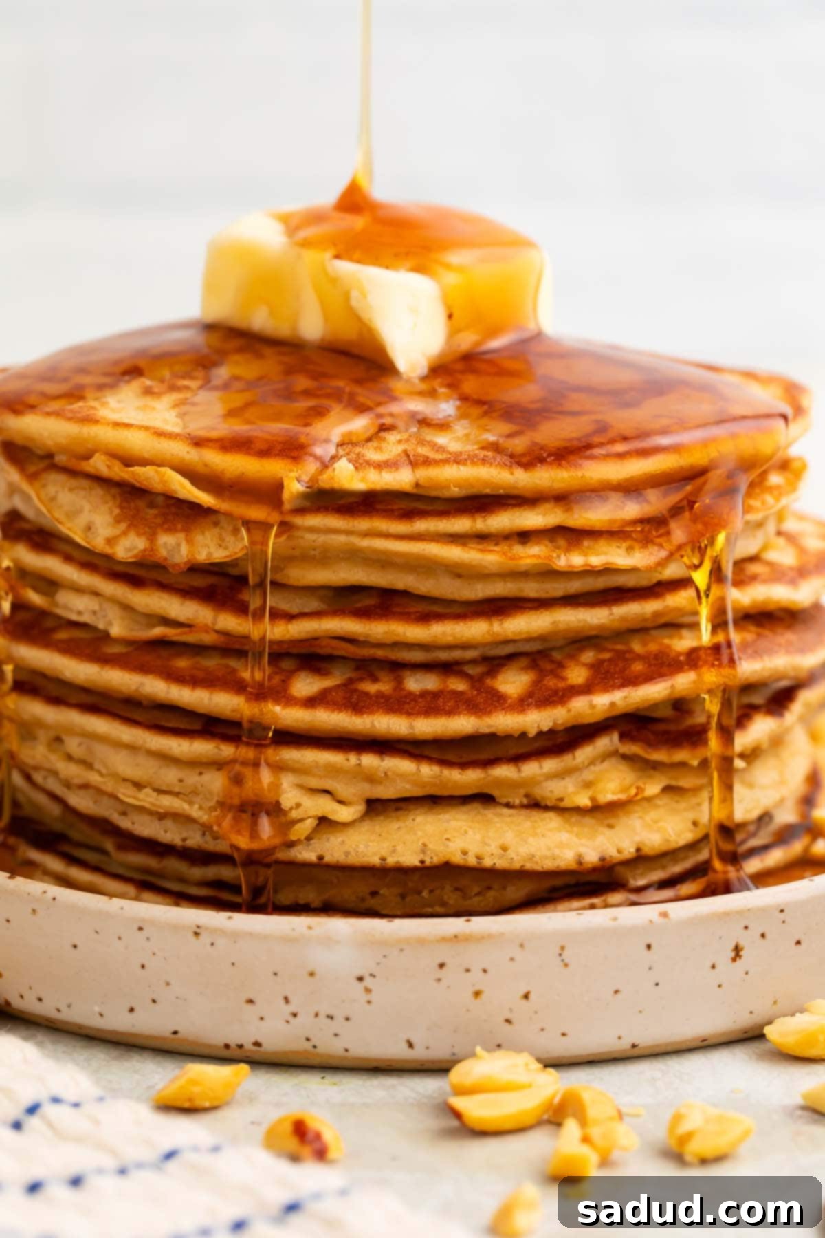 A tall stack of golden brown, fluffy peanut butter pancakes, generously topped with a melting pat of butter and a cascading stream of golden maple syrup, ready to be enjoyed.