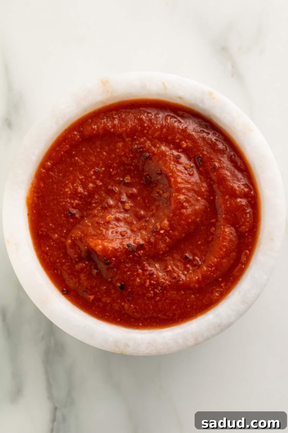 Top-down view of a small white bowl of deep red taco sauce on a marble countertop, garnished with a sprig of cilantro.