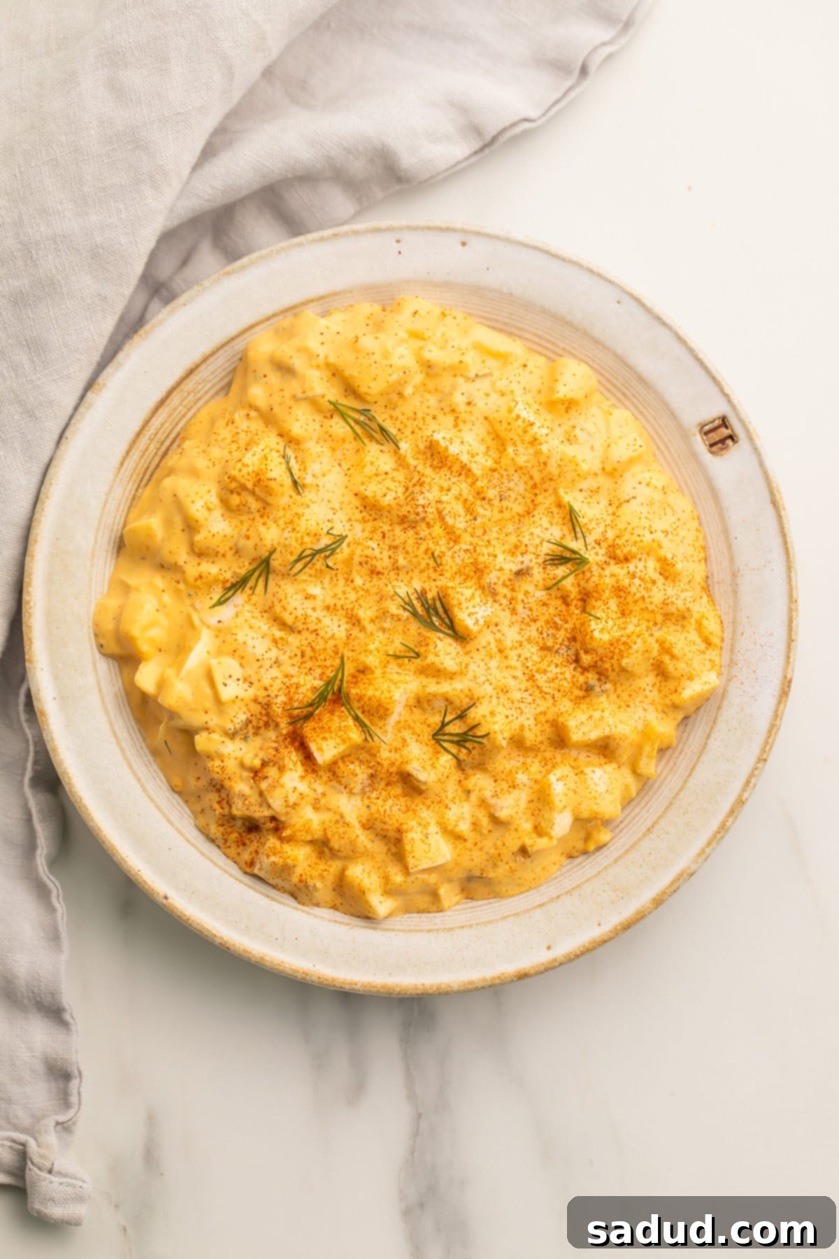 Overhead view of a bowl of creamy deviled egg salad on a neutral table with a cloth napkin and fresh parsley.