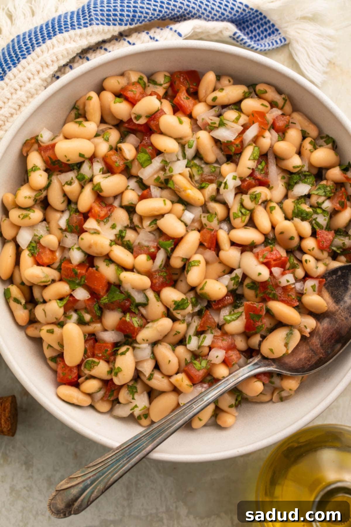 A large white bowl of white bean salad with onions, tomatoes, parsley, and a light vinaigrette, captured from a slightly elevated angle.