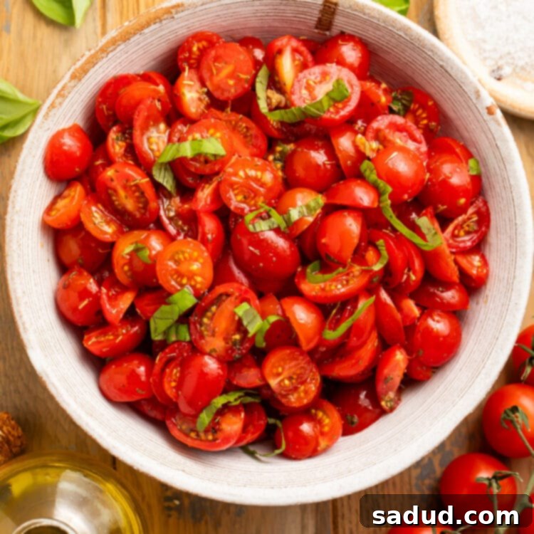 A large white bowl sits on a wooden table. In the bowl is a pile of sliced cherry tomatoes and strips of green fresh basil.