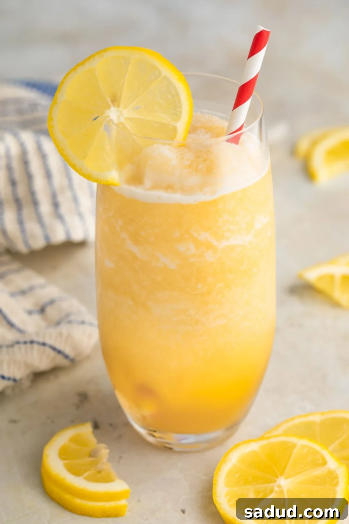 Effortless Frozen Lemonade 2 A tall glass filled with yellow frozen lemonade and garnished with a lemon coin and a red & white striped straw.