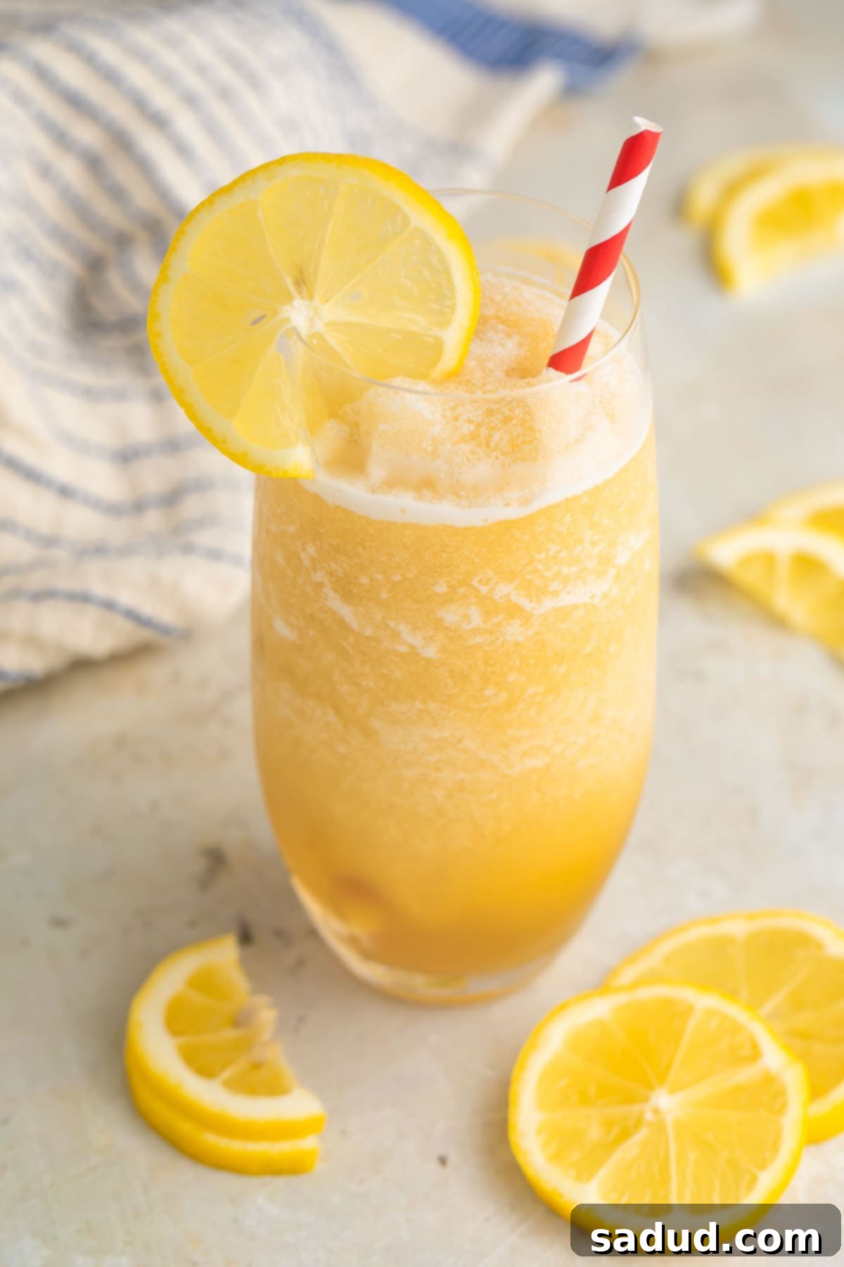 Effortless Frozen Lemonade 3 A tall glass filled with yellow frozen lemonade and garnished with a lemon coin and a red & white striped straw.