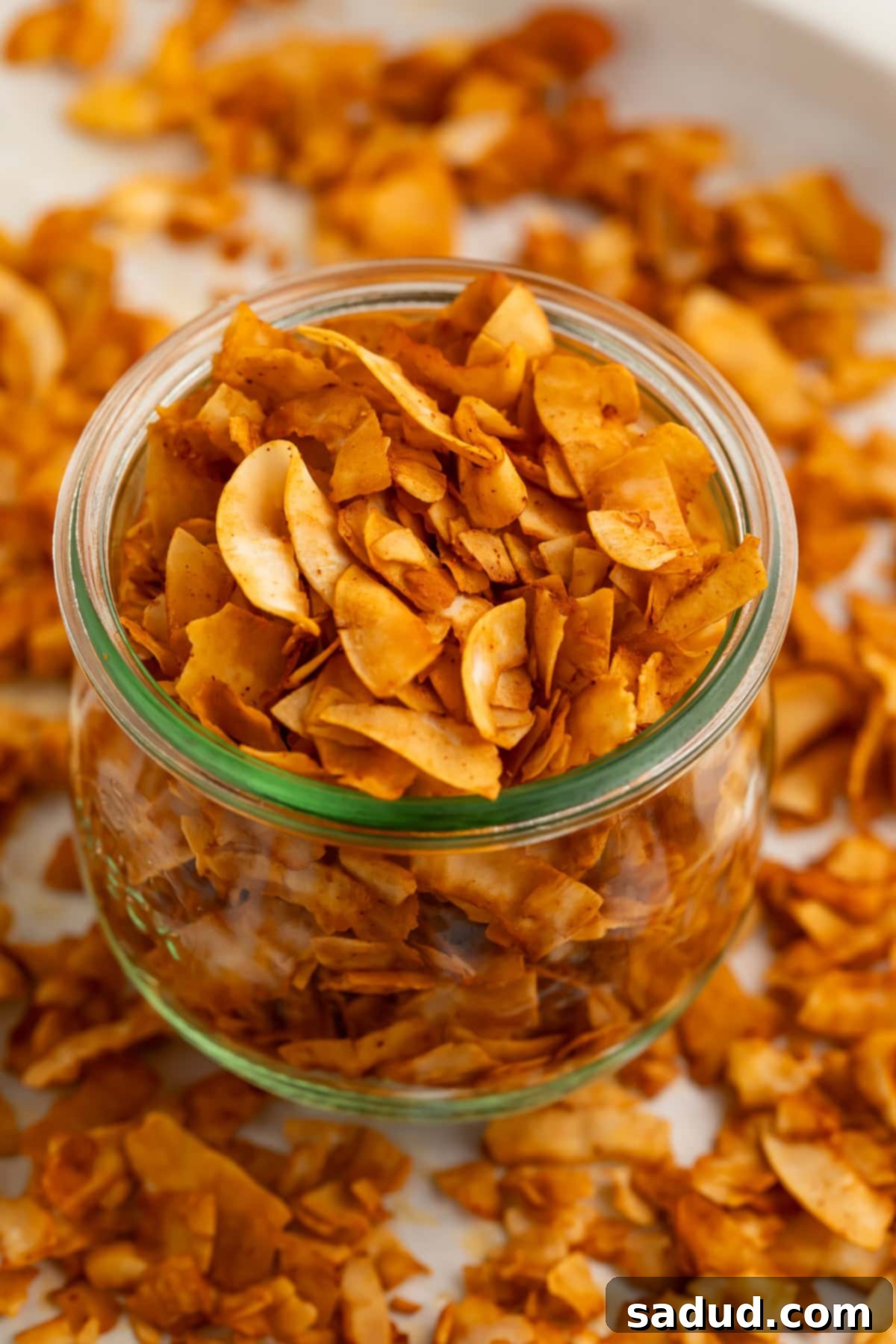 A glass jar of crispy coconut bacon chips on a table surrounded by coconut bacon.