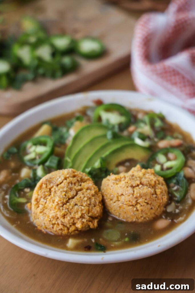 A steaming bowl of creamy vegan white bean chili, garnished with fresh cilantro and served alongside golden-brown, fluffy cornbread biscuits on a rustic wooden table.