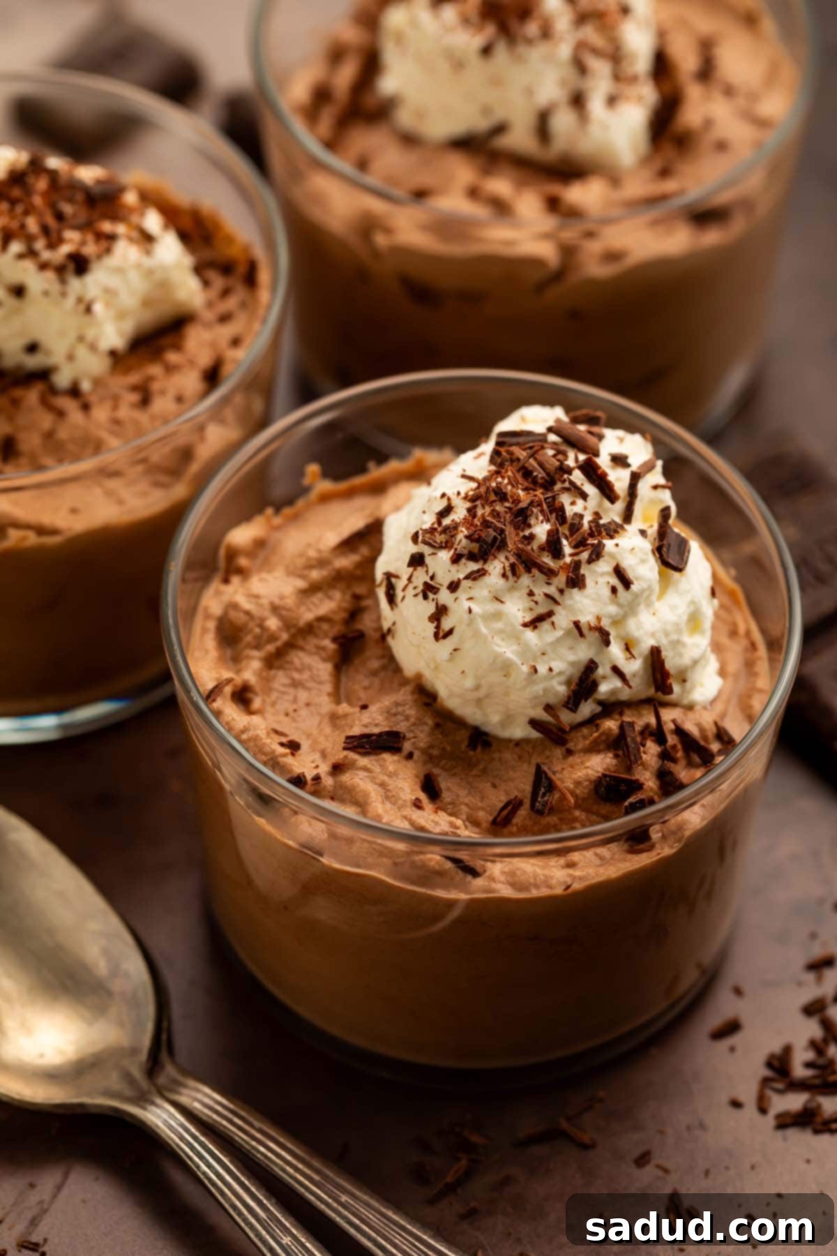 Three elegant glass cups of keto chocolate mousse, artfully topped with fluffy keto whipped cream and delicate chocolate shavings. The image highlights the rich, creamy texture and luxurious presentation of this low-carb dessert.