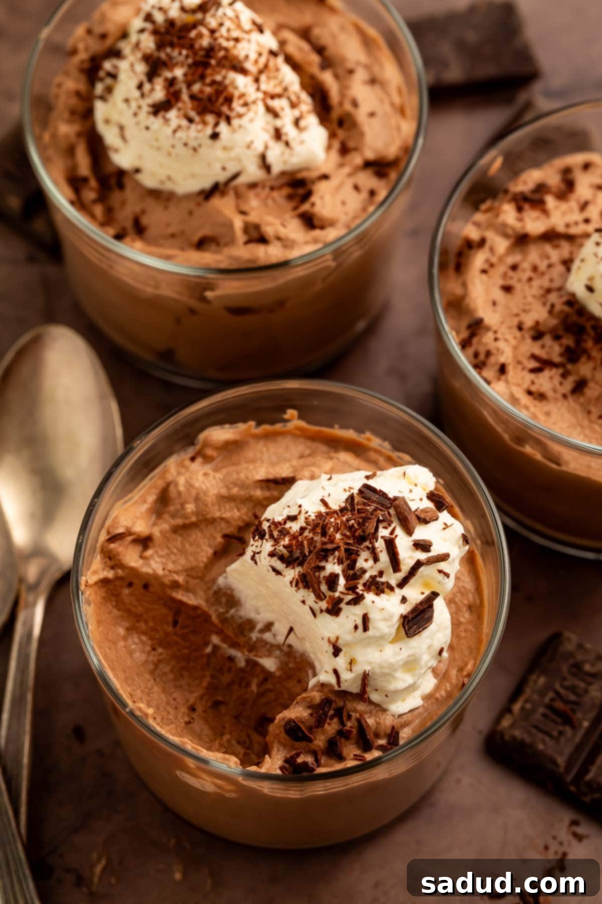 Close-up shot of three glass cups of keto chocolate mousse, artfully topped with fluffy keto whipped cream and delicate chocolate shavings, ready to be served. The focus is on the rich texture and appealing presentation.