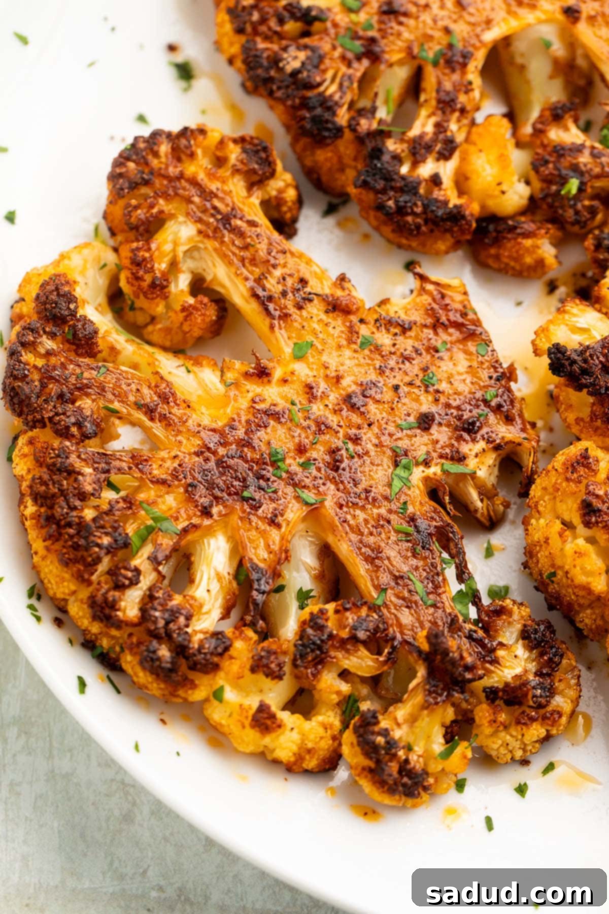 Two perfectly roasted cauliflower steaks, golden brown and garnished with fresh parsley, sit on a white plate ready to be served. The rich seasoning is clearly visible.