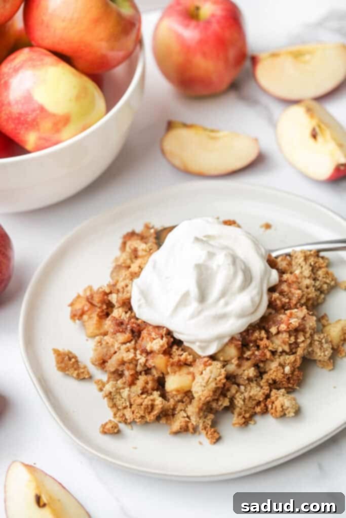 A serving of warm vegan apple crisp on a white plate, with a bowl of fresh red apples in the blurred background. The crisp has a golden, crumbly topping and a visible apple filling.