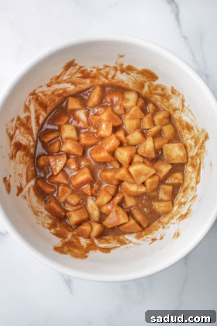 Chopped red apples being coated in a creamy cashew butter and maple syrup mixture in a large bowl.