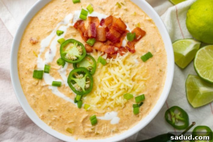 A close-up shot of a full bowl of creamy keto chicken white chili, garnished generously with crispy bacon pieces, vibrant green jalapeno slices, and fresh lime wedges on the side, emphasizing its rich texture and delicious toppings.