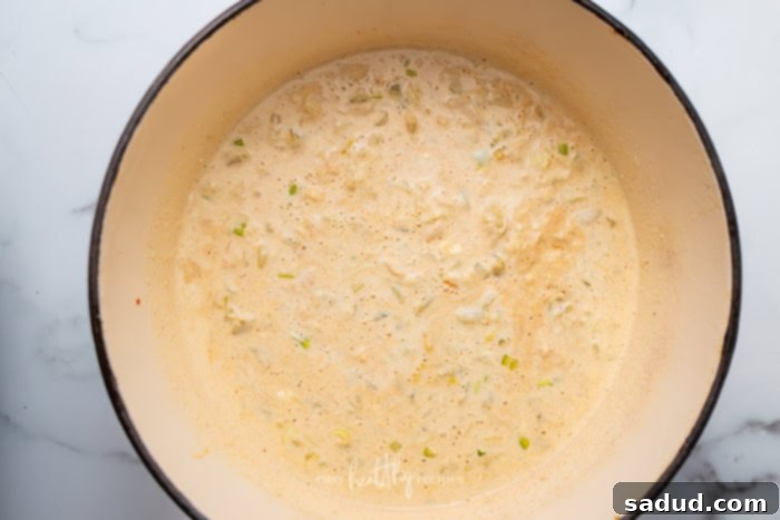 A creamy chili base with chicken broth, heavy cream, cream cheese, and sriracha simmering in a pot, with shredded chicken, Monterey Jack cheese, and diced green chilis added and being stirred.