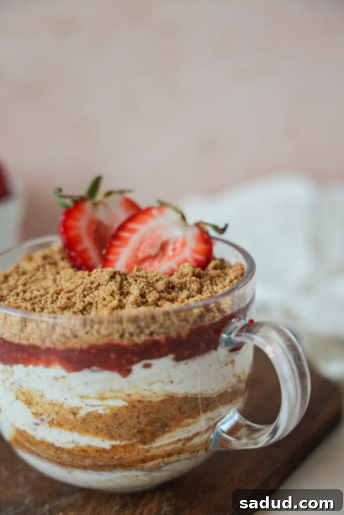 A close-up shot of a vegan strawberry cheesecake parfait in a clear glass, showcasing delicious layers of creamy white base, vibrant red strawberry jam, and a topping of golden-brown cookie crumbles.