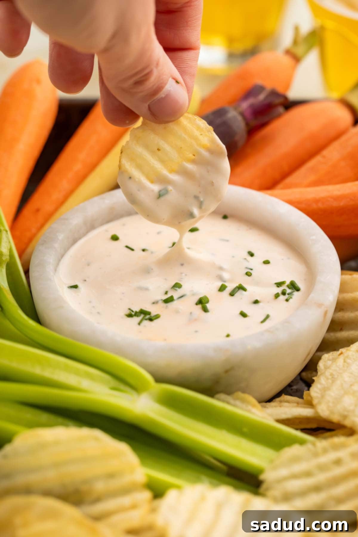A woman's hand holding a potato chip with ridges above a bowl of buffalo ranch dressing as dressing drips off the chip and back into the bowl.