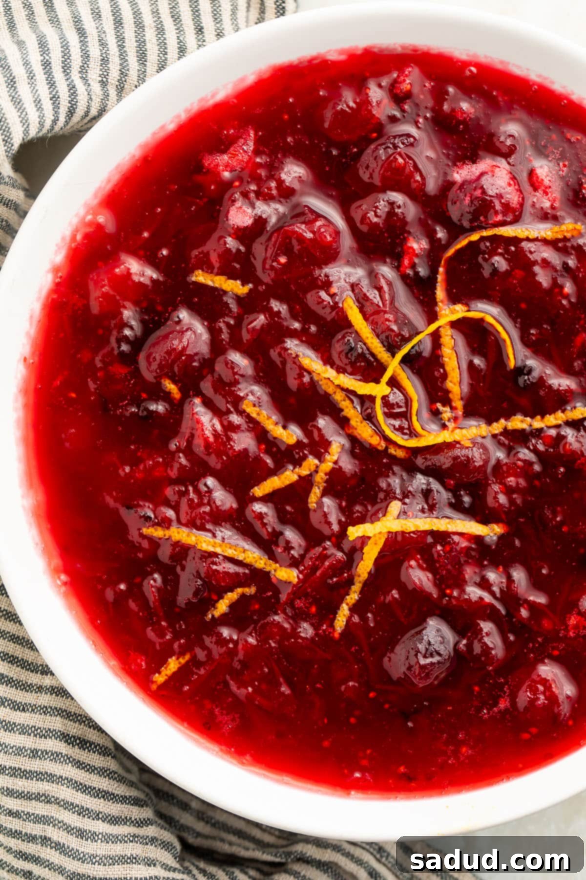 Close-up view of a white bowl filled with deep red homemade keto cranberry sauce, with whole cranberries throughout, garnished with vibrant orange zest.