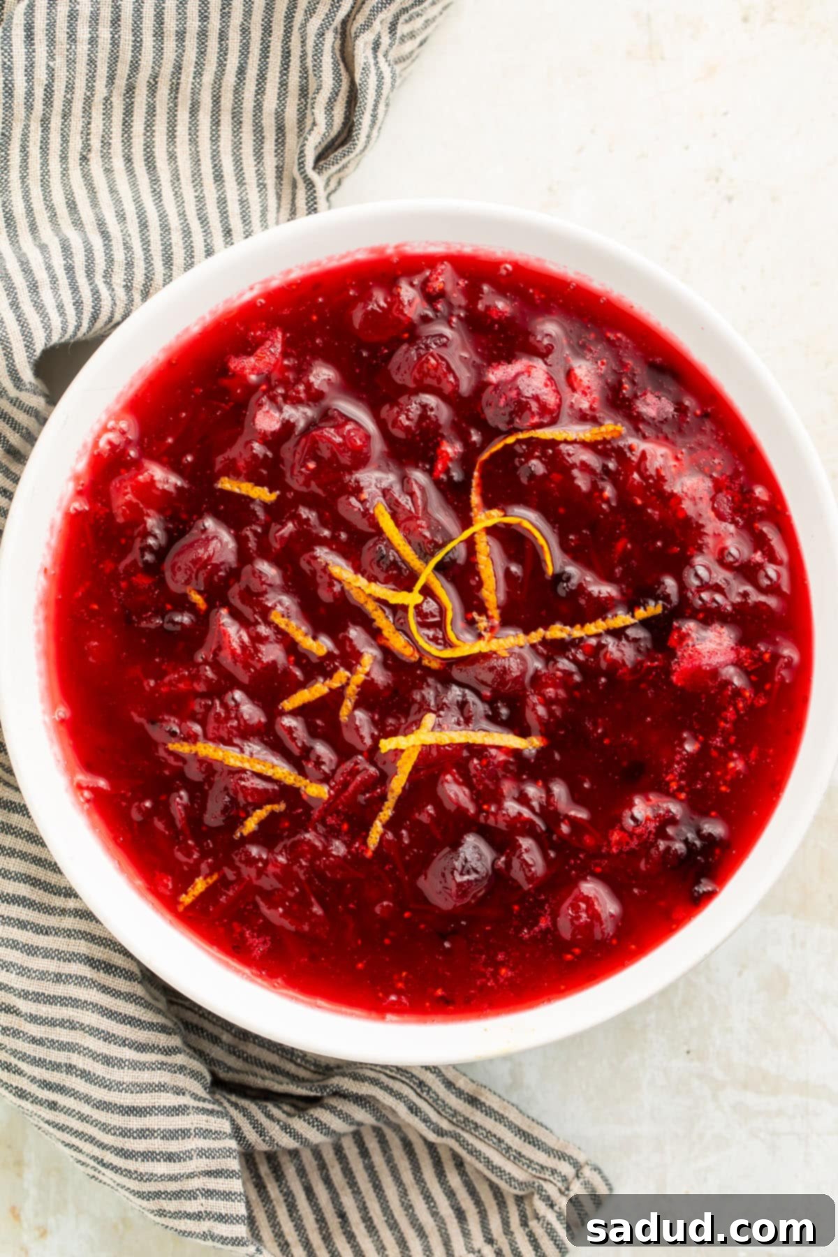 Top-down, overhead view of a white bowl filled with deep red homemade keto cranberry sauce, with whole cranberries throughout, garnished with fresh orange zest.