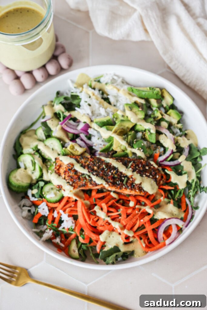 A close-up of a well-arranged Salmon Nourish Bowl with cooked rice, a piece of flaky salmon, various colorful vegetables like cucumber and carrots, and a generous drizzle of golden tahini dressing.