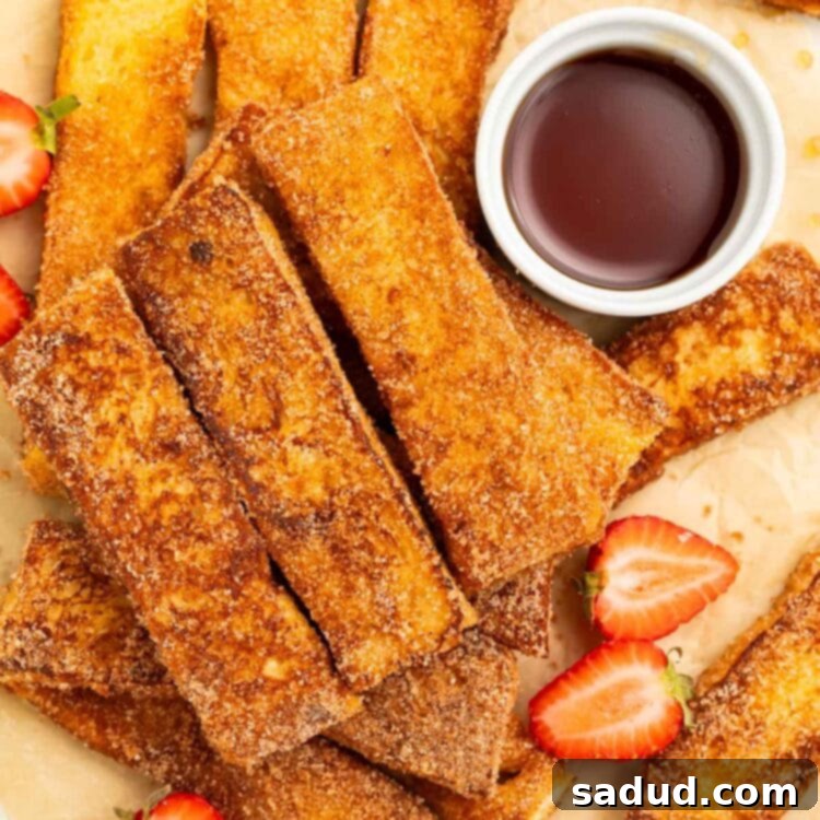 Keto French toast sticks arranged in a pile on a sheet of parchment paper. A ramekin of keto syrup and slices of strawberries sit next to the french toast sticks.