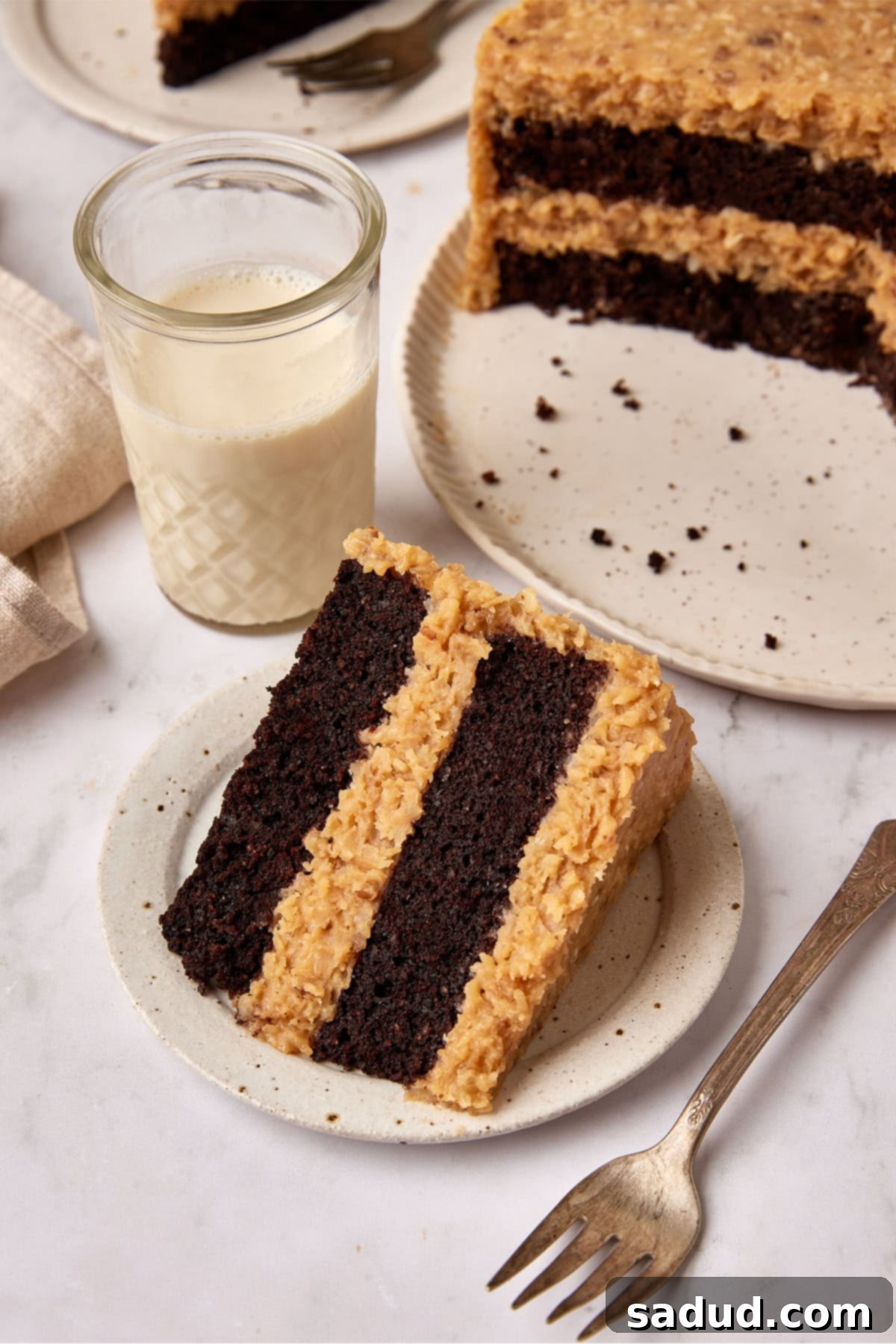 Slice of Keto German Chocolate cake on a small white plate with a fork, next to a glass of milk. A whole cake is visible in the soft background.