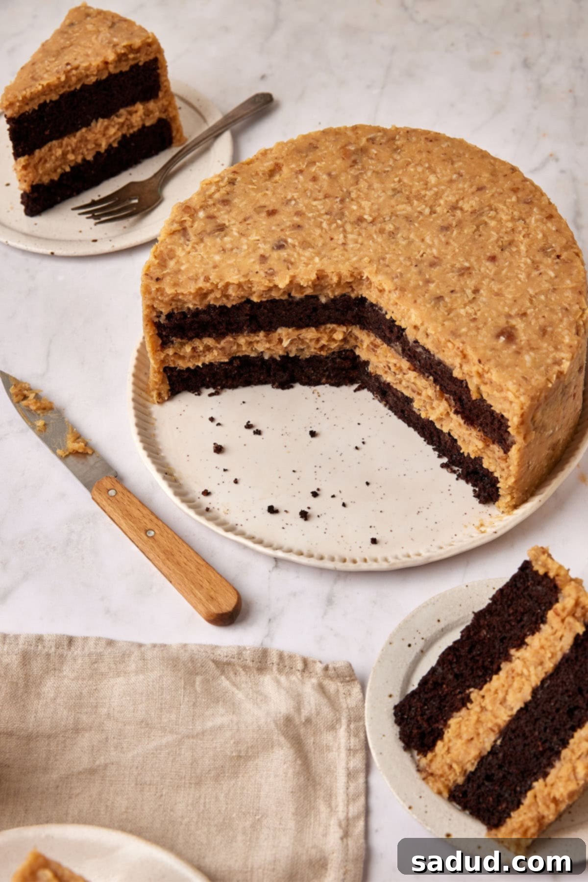 A 2/3 cut Keto German Chocolate Cake with slices on plates, a napkin, and a knife surrounding it on a kitchen countertop, showcasing its rich layers and frosting.
