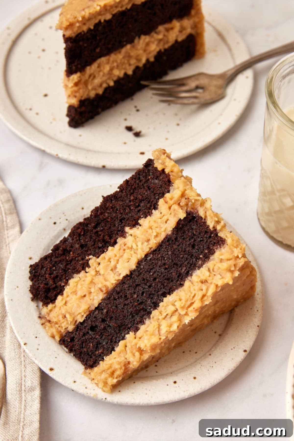 A thick, perfectly sliced piece of Keto German Chocolate Cake on a white plate, with another slice and a fork in the soft focus background, highlighting the rich frosting and cake layers.