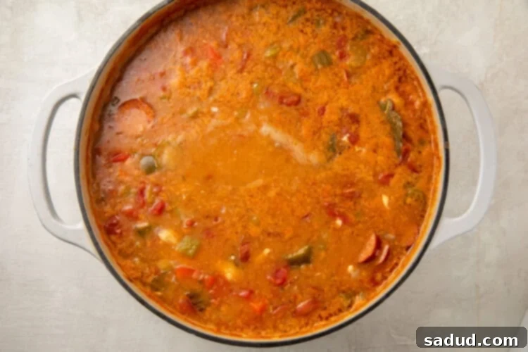 A large pot filled with a simmering keto gumbo, showing broth, diced tomatoes, okra, and shrimp beginning to cook, with bay leaves visible.