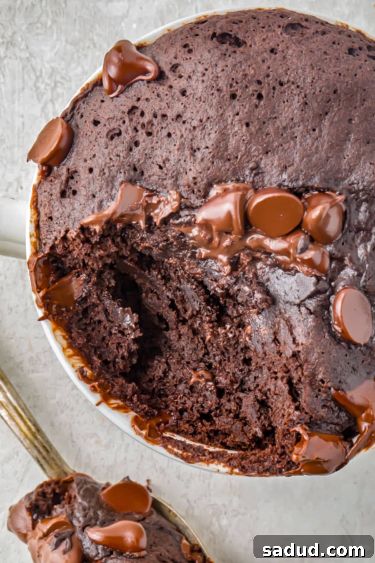A close-up, top-down view of a rich chocolate gluten-free mug cake in a white mug, with a generous spoonful of cake already scooped out and resting on the tarnished spoon, showcasing its moist texture.