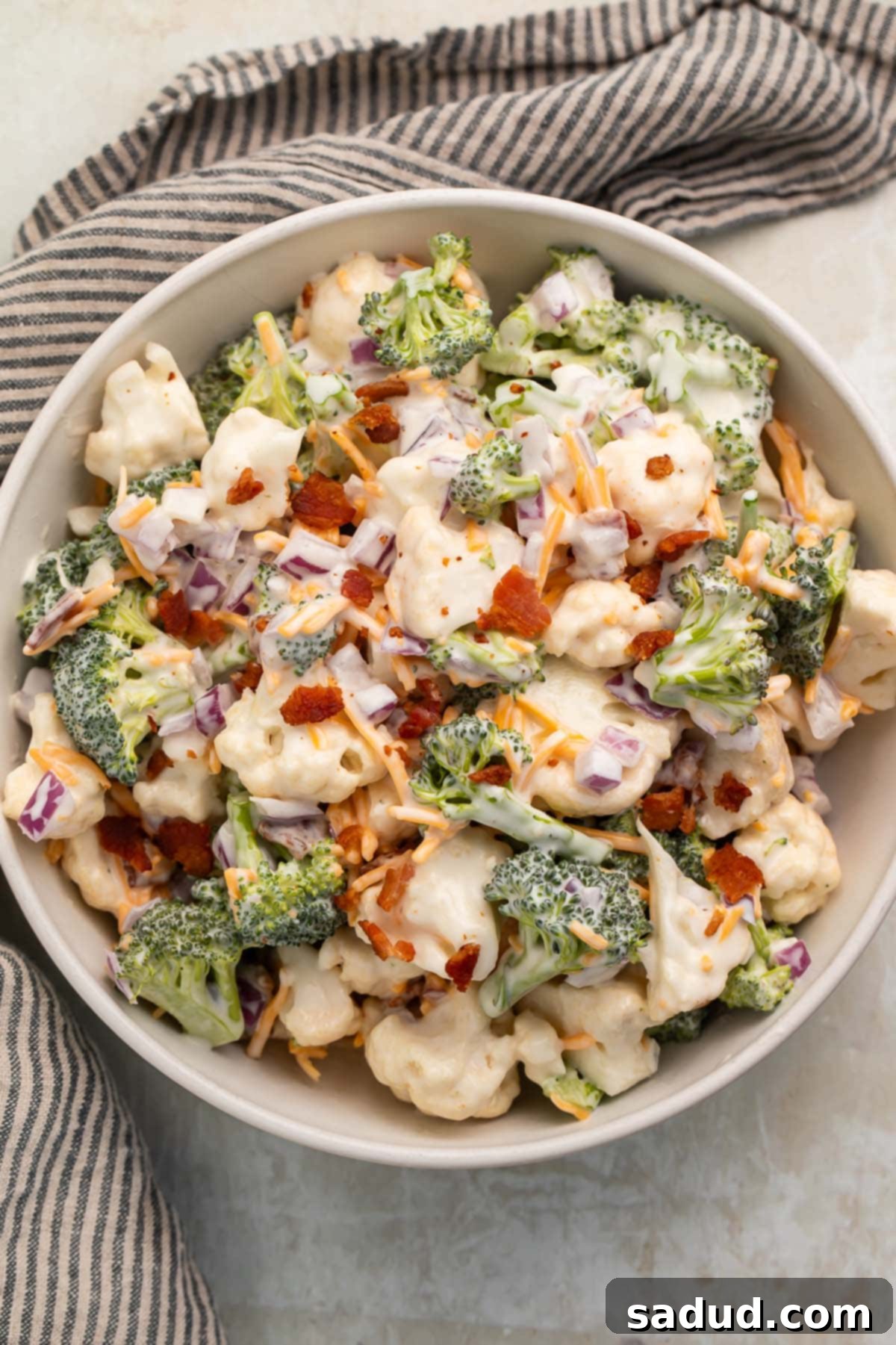 A close-up view of a large white bowl filled with creamy Amish broccoli salad, showcasing the texture of the broccoli and cauliflower florets, crispy bacon pieces, and shredded cheddar cheese mixed in the rich dressing.