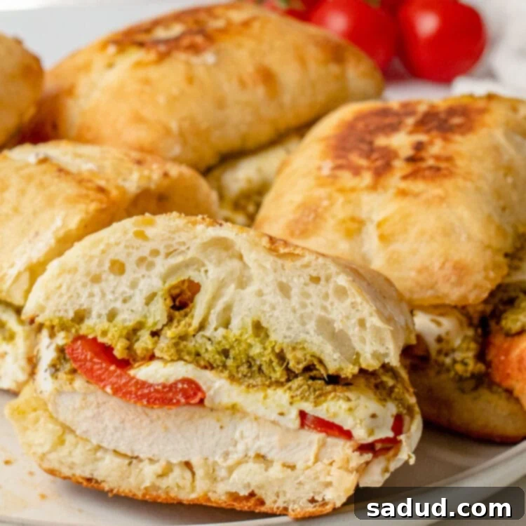 Zesty Pesto Chicken Sandwich 4 Cross section of a chicken pesto sandwich resting on the lip of a plate, with other sandwiches in the background.