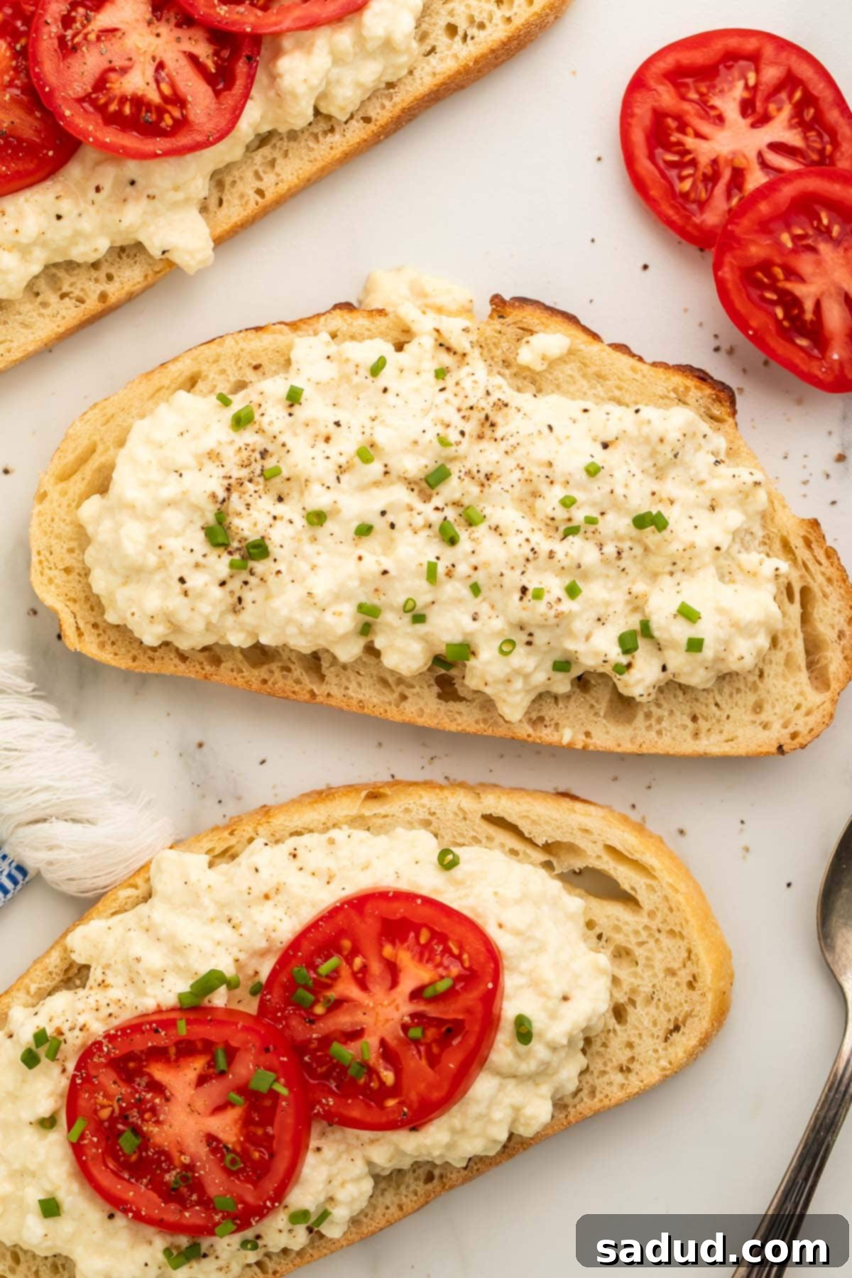 Homemade vegan cottage cheese spread generously on slices of crusty French bread, adorned with vibrant red tomato slices and a sprinkle of fresh herbs.