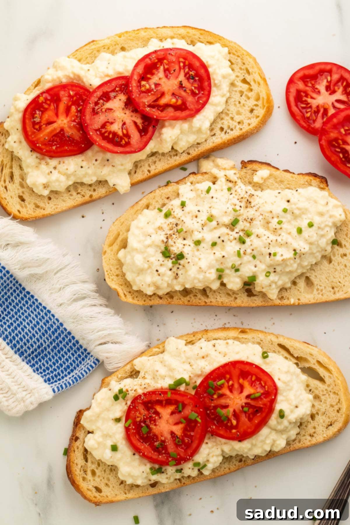 Close-up of homemade vegan cottage cheese spread on slices of French bread, garnished with vibrant red tomato slices and fresh green herbs, showcasing its creamy yet chunky texture.