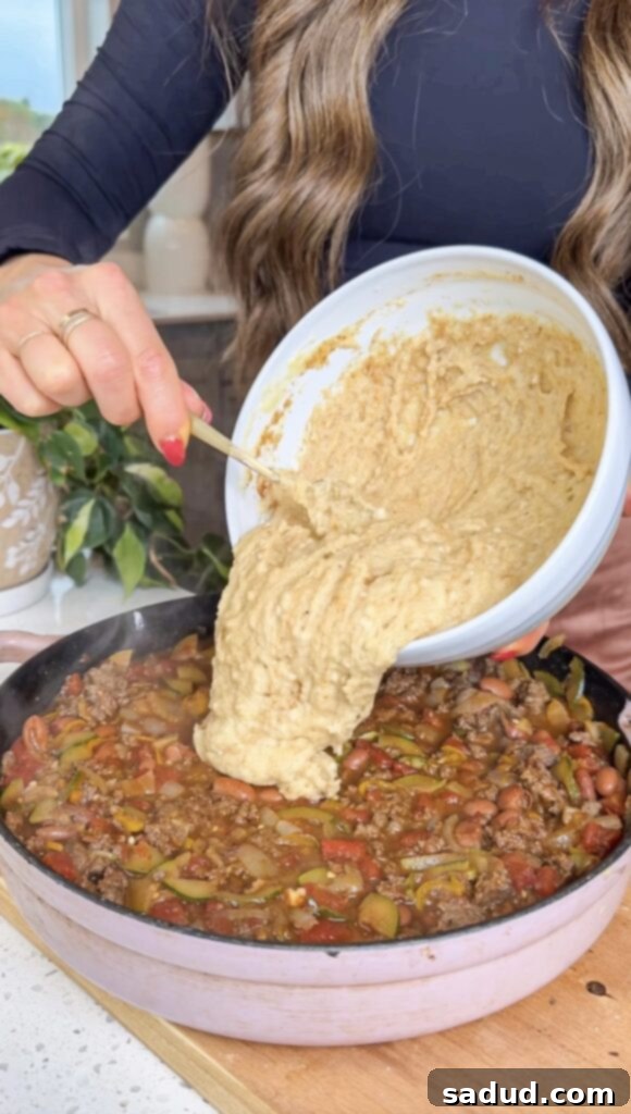 A close-up of the cornbread batter being poured over the chili in a casserole dish.
