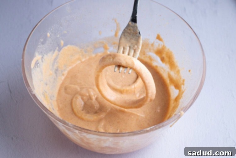 Coated onion ring held over batter in a large glass mixing bowl with a fork