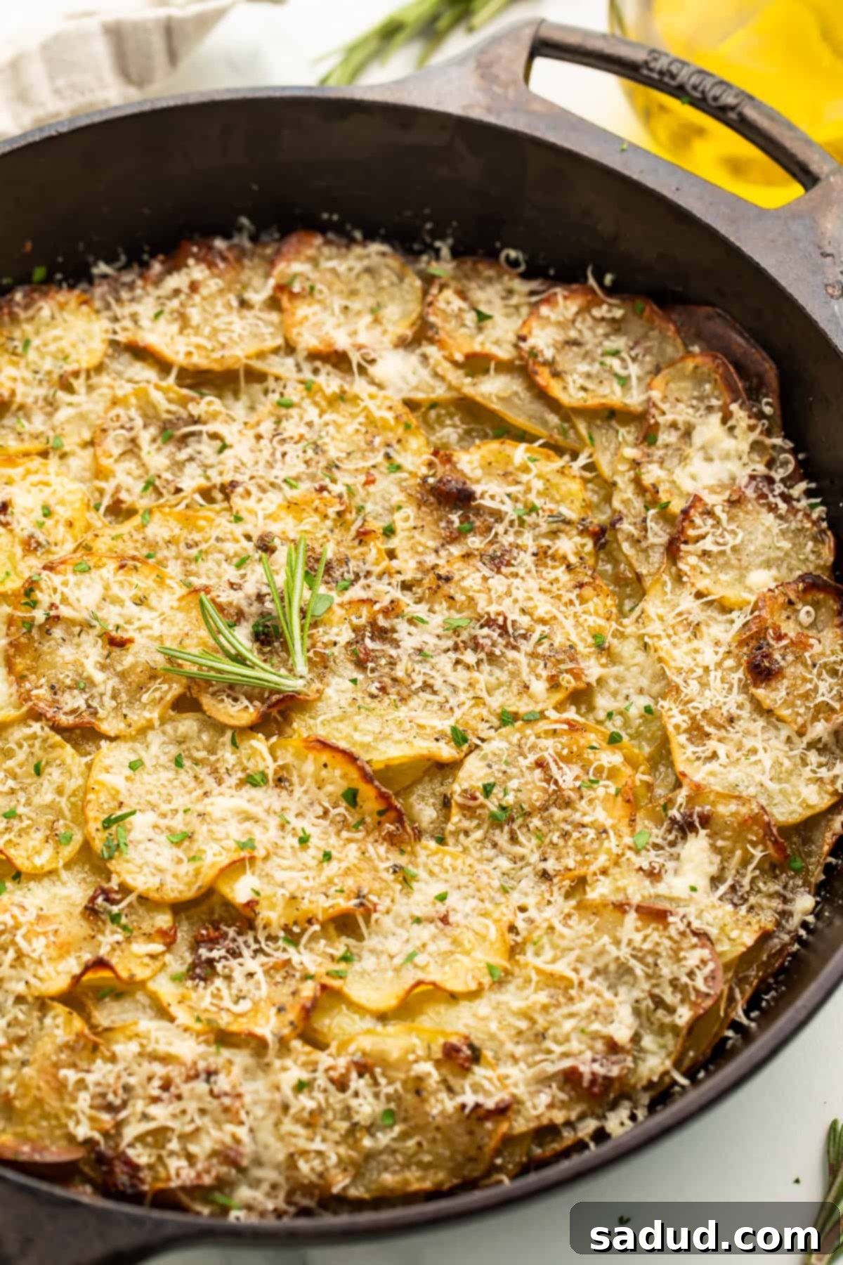 Close up image of a potato galette in a cast iron skillet, covered in melted grated cheese and garnished with a fresh rosemary tip, highlighting the crispy edges.