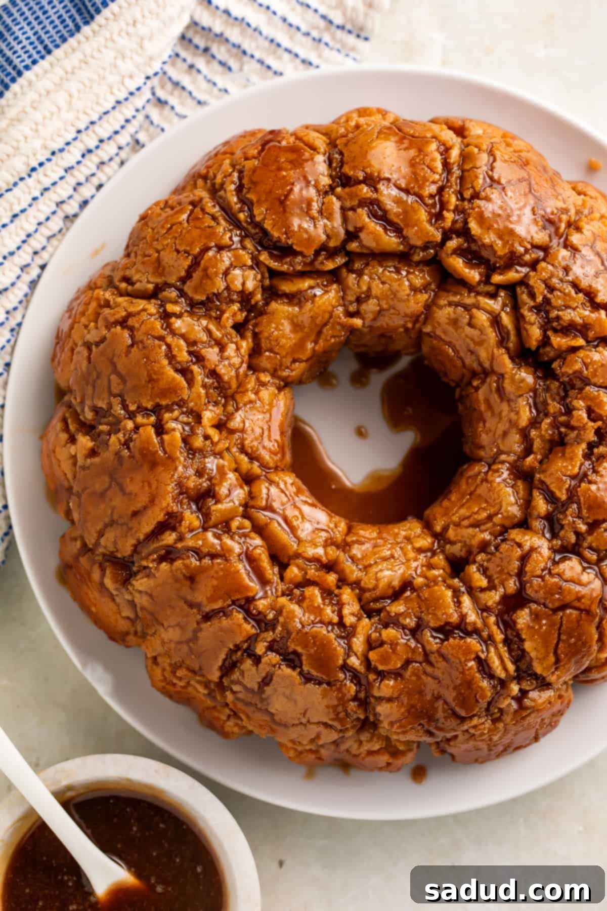 Overhead view of a monkey bread drizzled with caramel on a white plate. A white bowl of caramel with a white spoon sits off to one side.