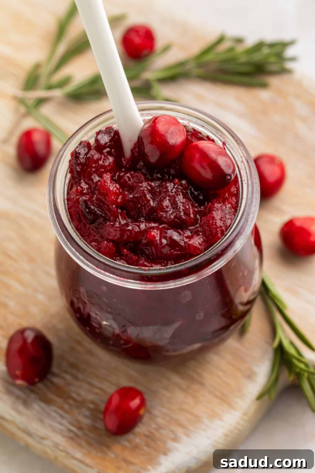 Jar of open cranberry jam with a white spoon on a wooden board scattered with rosemary sprigs and fresh cranberries.