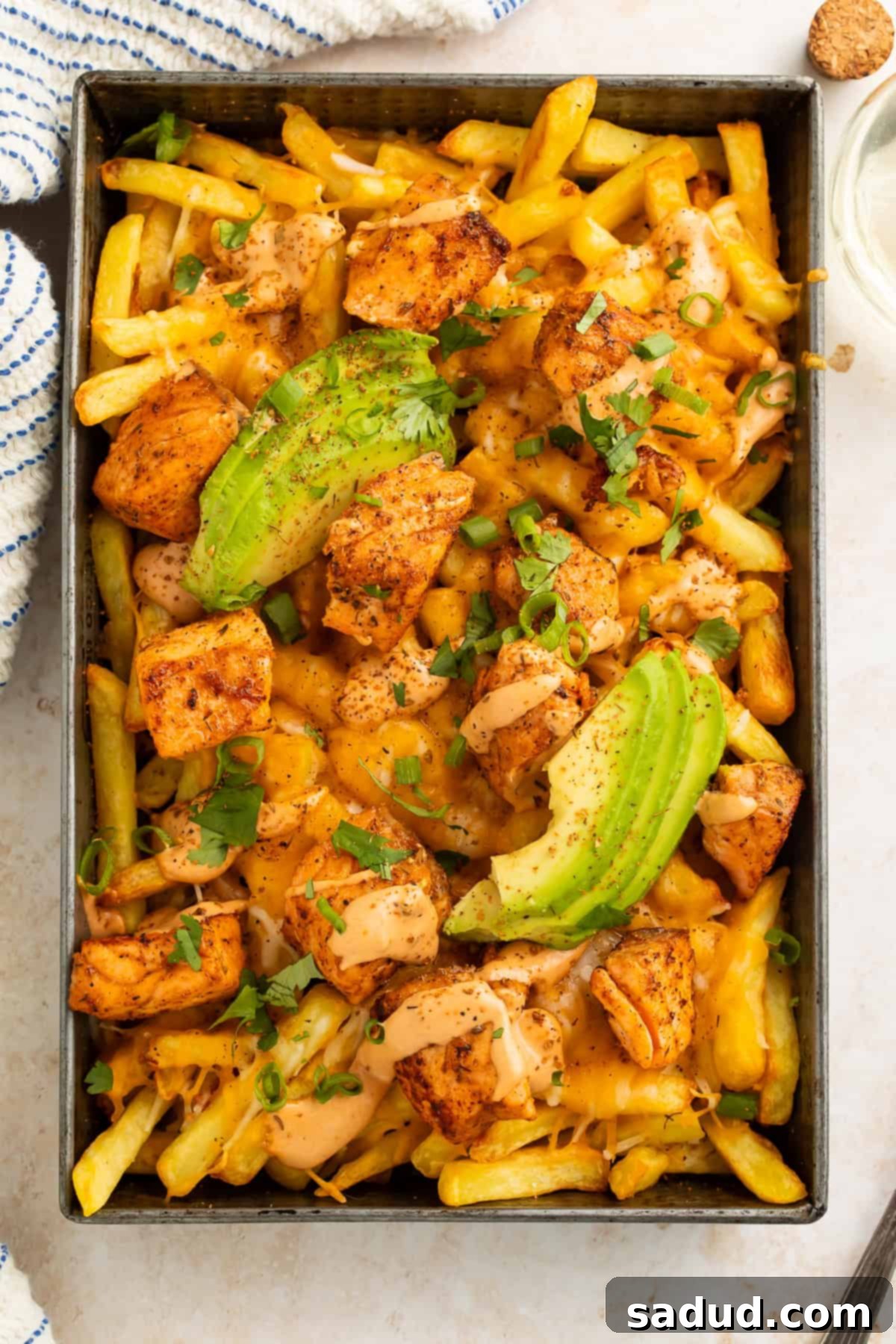 Metal tray of cooked fries topped with blackened salmon, creamy sauce and sliced avocado. 